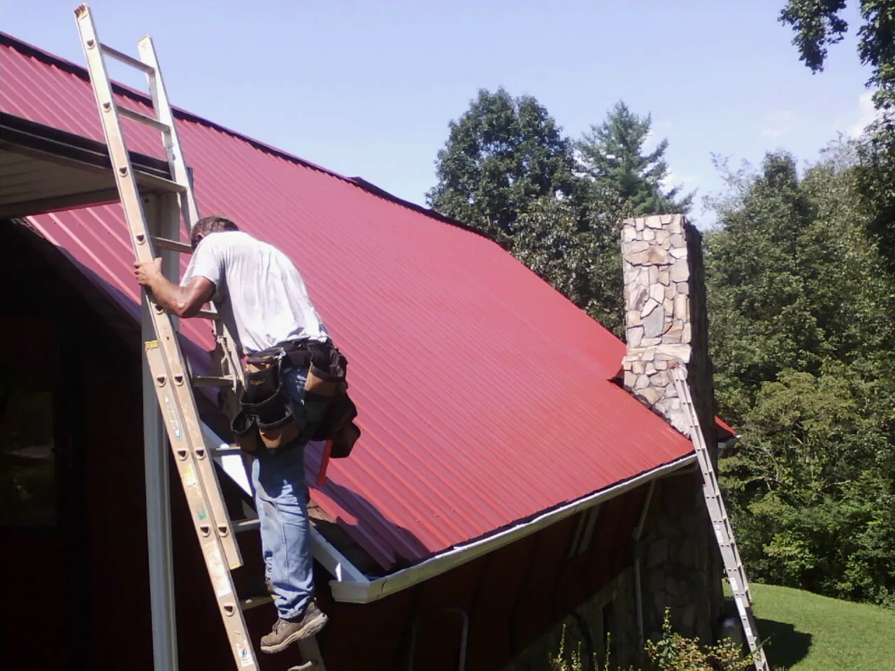 Roofer on a ladder beside a red metal roof and stone chimney — Soffit & Fascia Repair by Roofing Brothers in Happy Jack